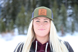 Model wears a forest green trucker hat. There is a leather patch sewn on with peace fingers, a heart, and MT engraved. A frozen lake and mountains are featured in the background.