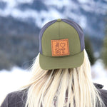Model wears a forest green trucker hat backwards. There is a leather patch sewn on with peace fingers, a heart, and MT engraved. A frozen lake and mountains are featured in the background.